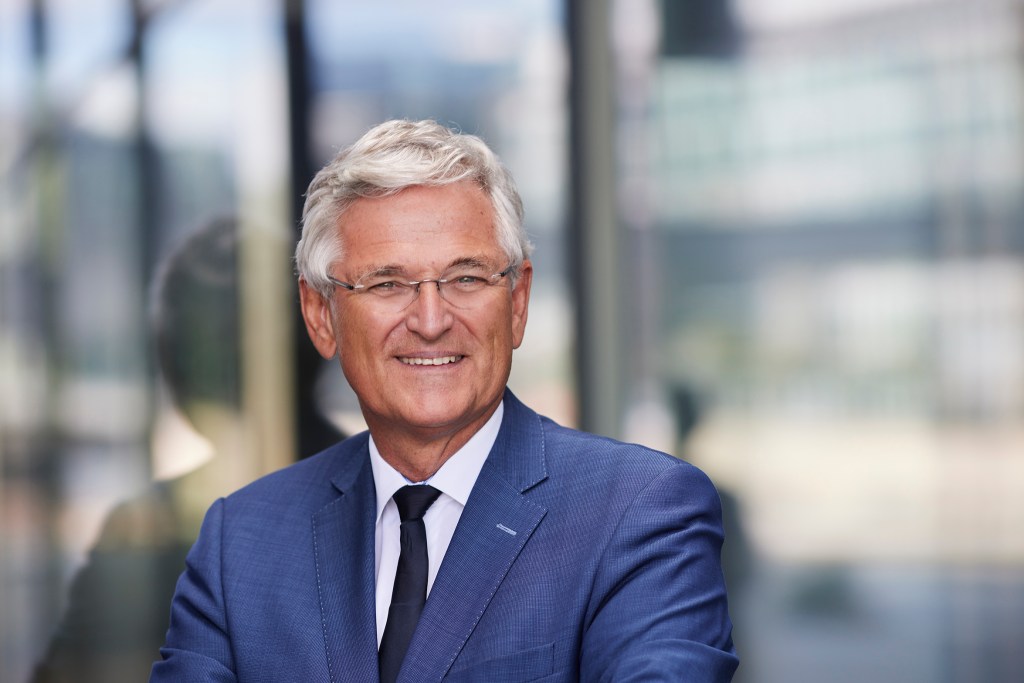 Ein älterer Mann mit grauen Haaren und Brille, der in einem blauen Anzug lächelt, steht vor einer modernen Glasarchitektur.